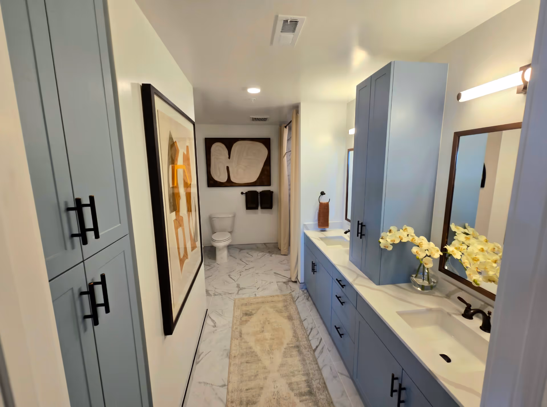 A modern bathroom with light blue cabinets, a double sink vanity with two mirrors, a toilet at the far end, abstract artwork on the walls, and a beige patterned rug on the marble floor.