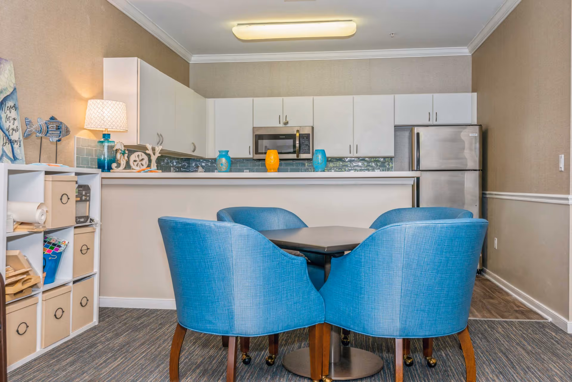 A small dining area with a square table surrounded by four blue upholstered chairs. Behind the dining area is a kitchen with white cabinets, a stainless steel refrigerator, microwave, and a countertop decorated with colorful vases and small decorative items. To the left, there is a white shelving unit with storage bins and various items. The walls are beige with white trim, and the floor has a carpeted area transitioning to a wood floor in the kitchen.