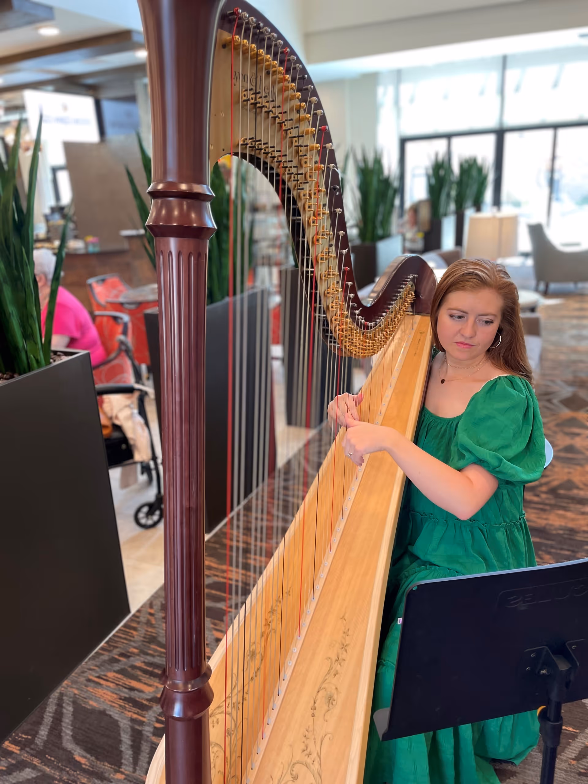 A woman in a green dress playing a large wooden harp in a well-lit indoor space with plants and seating areas in the background.