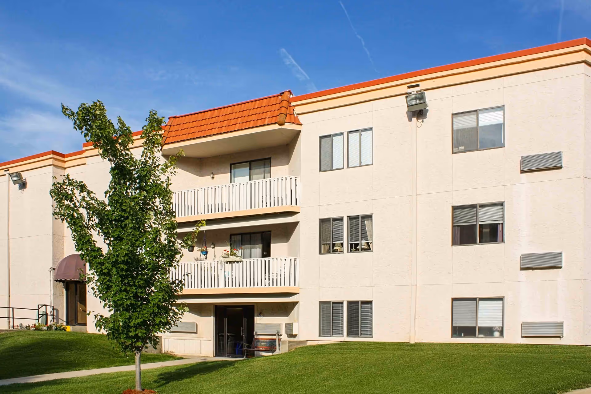Exterior view of a three-story senior living facility building with beige walls and orange roof tiles. The building features balconies with white railings, several windows, a green lawn, and a small tree in the foreground under a clear blue sky.