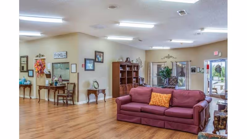A spacious senior living common room with a maroon sofa, wooden floors, bookshelves, side tables and seating near windows and an exit door.