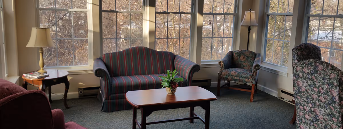 A cozy living room with large windows letting in natural light. The room features a striped loveseat, floral armchairs, a wooden coffee table with a small potted plant, and two lamps on side tables. The carpet is blue, and the walls are light-colored.