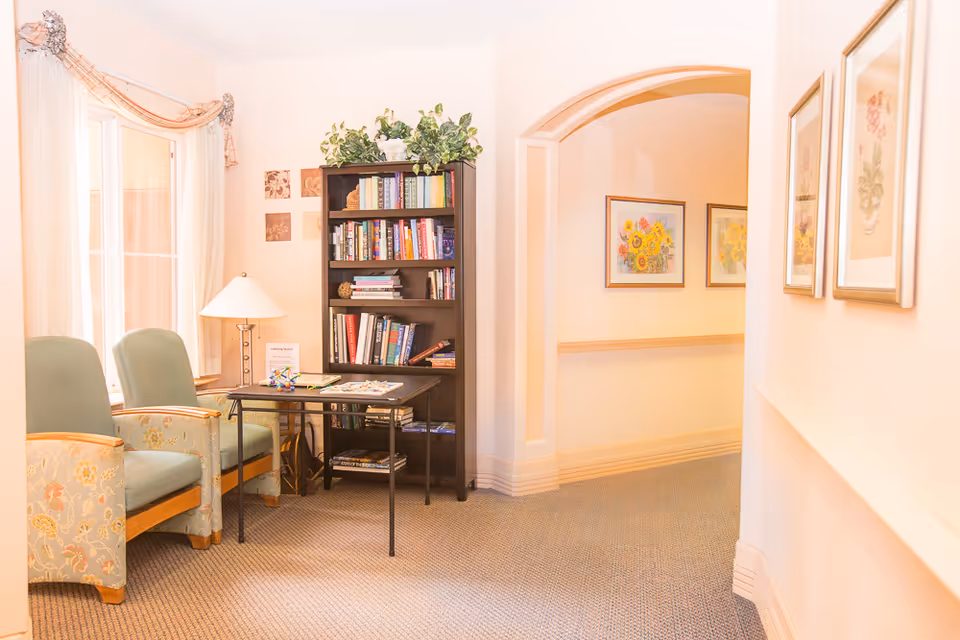 Sunlit seating nook with two upholstered chairs, a lamp, a small table and a bookshelf in a hallway.