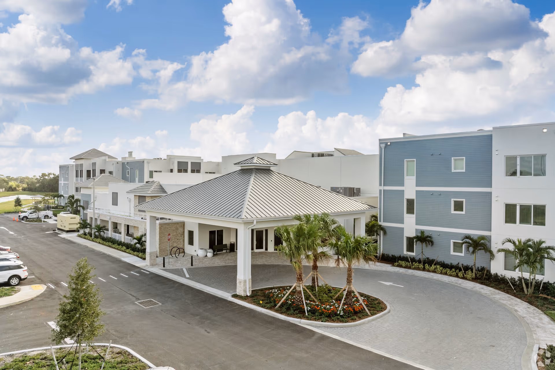 Exterior view of AVIVA Port St. Lucie West senior living facility showing a covered entrance with a metal roof, surrounded by palm trees and landscaped flower beds. The building has multiple stories with white and blue-gray walls, and there is a parking area with cars on the left side. The sky is partly cloudy.