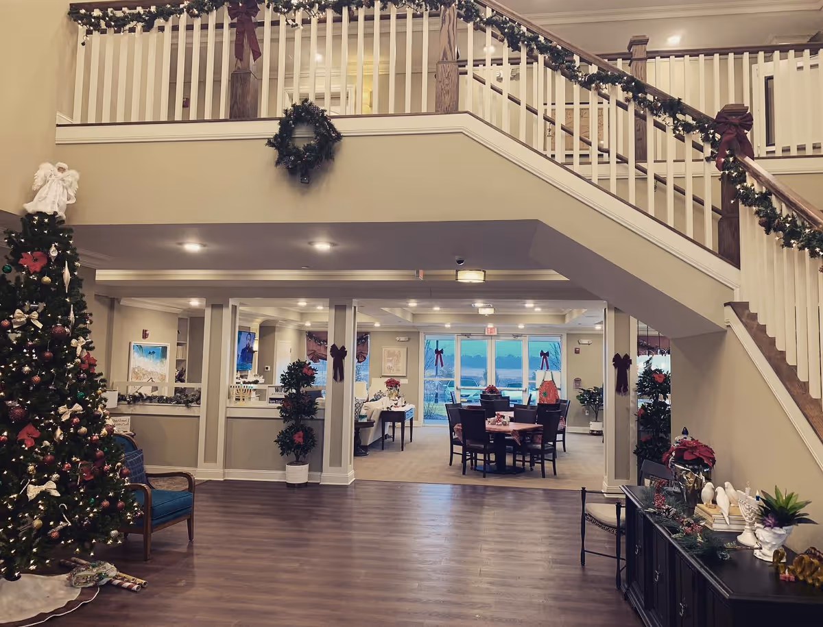 Interior view of a senior living facility lobby decorated for Christmas with a large Christmas tree, garlands with bows on the staircase railing, wreaths on the walls, and festive decorations on furniture. There is a seating area with chairs and tables in the background near large windows.
