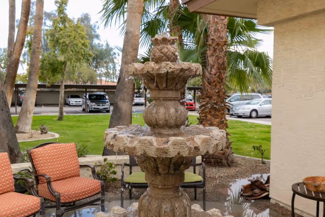 A tiered stone fountain in a courtyard with patio chairs, palm trees, and parked cars visible in the background.