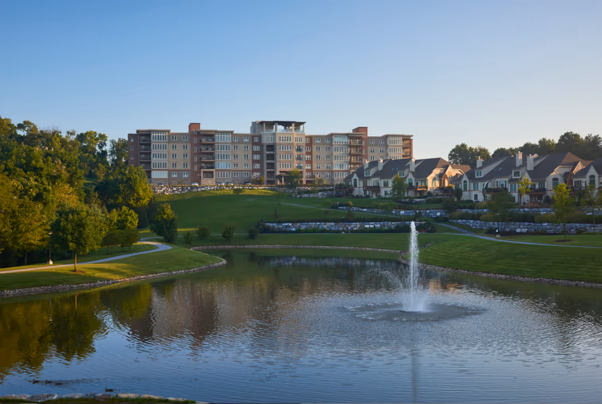 A scenic view of The Vistas at Willow Valley Communities featuring a large pond with a water fountain in the foreground, surrounded by green lawns and trees. In the background, there is a multi-story residential building and several smaller townhouse-style homes under a clear blue sky.