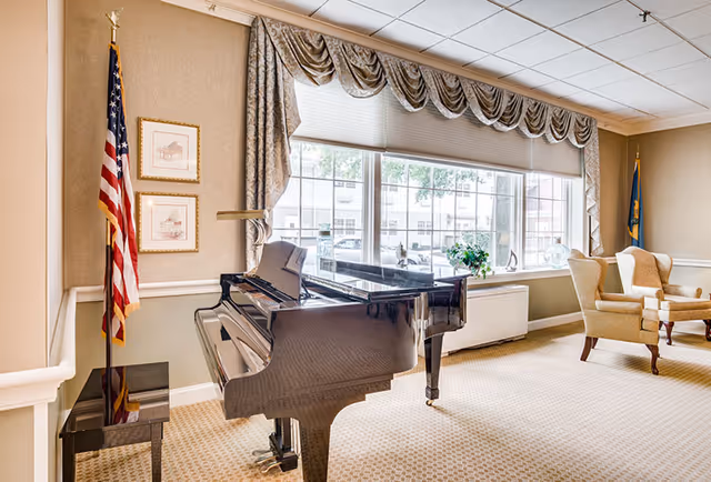 A bright room with a black grand piano and a matching piano bench near a large window with patterned curtains. Two beige upholstered armchairs are positioned near the window. An American flag stands in the corner next to two framed pictures on the wall. The room has beige carpet and light-colored walls.