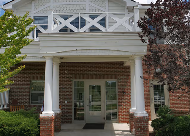 Entrance of a brick building with white pillars supporting a balcony above. The entrance has double glass doors with windows on either side. There are green bushes and trees around the entrance.