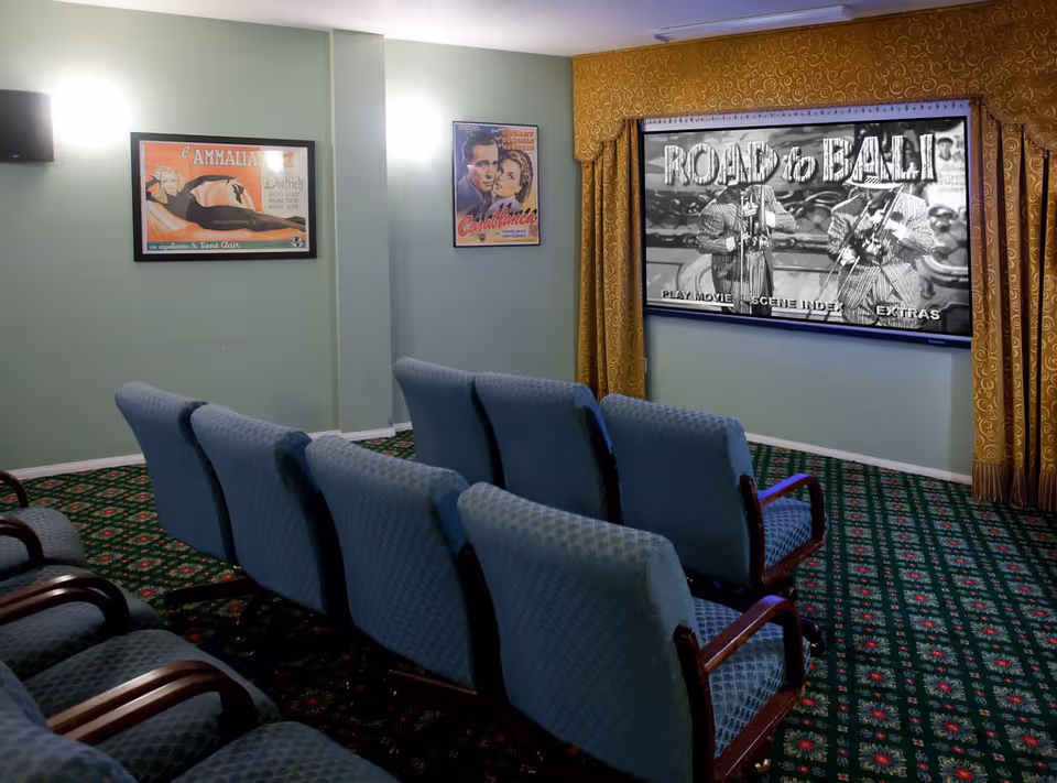 A small movie theater room with two rows of blue cushioned chairs facing a large screen showing a black and white movie titled 'Road to Bali'. The room has green walls with two framed vintage movie posters and patterned green carpet. Gold curtains frame the screen.