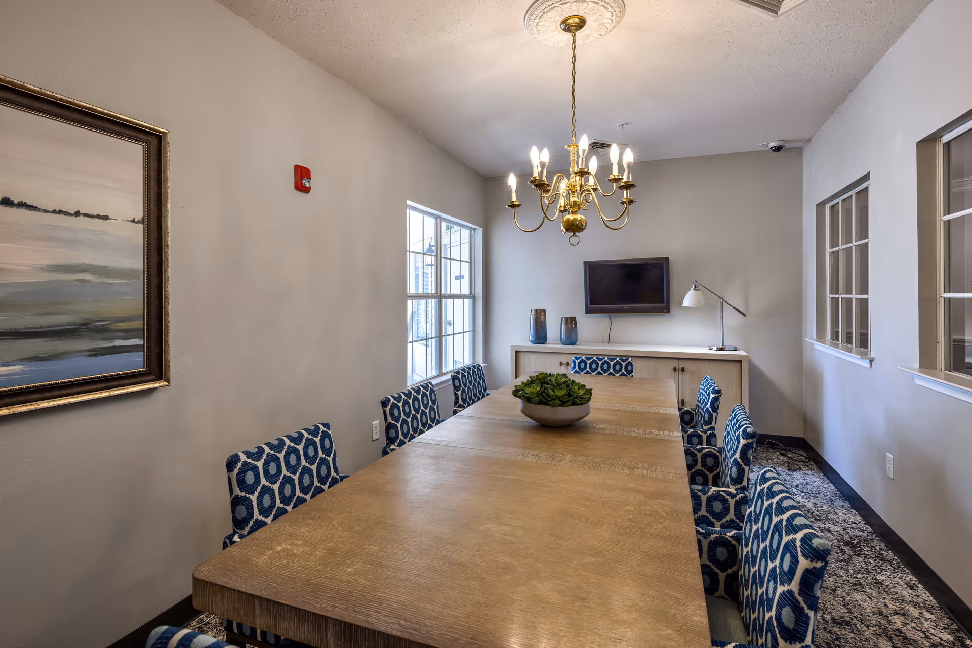 Conference-style dining room with a long wooden table surrounded by patterned blue chairs, a chandelier overhead, a wall-mounted TV and windows.
