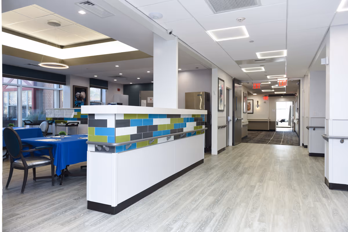Interior view of a senior living facility hallway with a reception desk featuring colorful tile accents. To the left, there is a dining area with tables covered in blue tablecloths and chairs. The hallway has light wood flooring, white walls, and modern ceiling lights with exit signs visible in the distance.