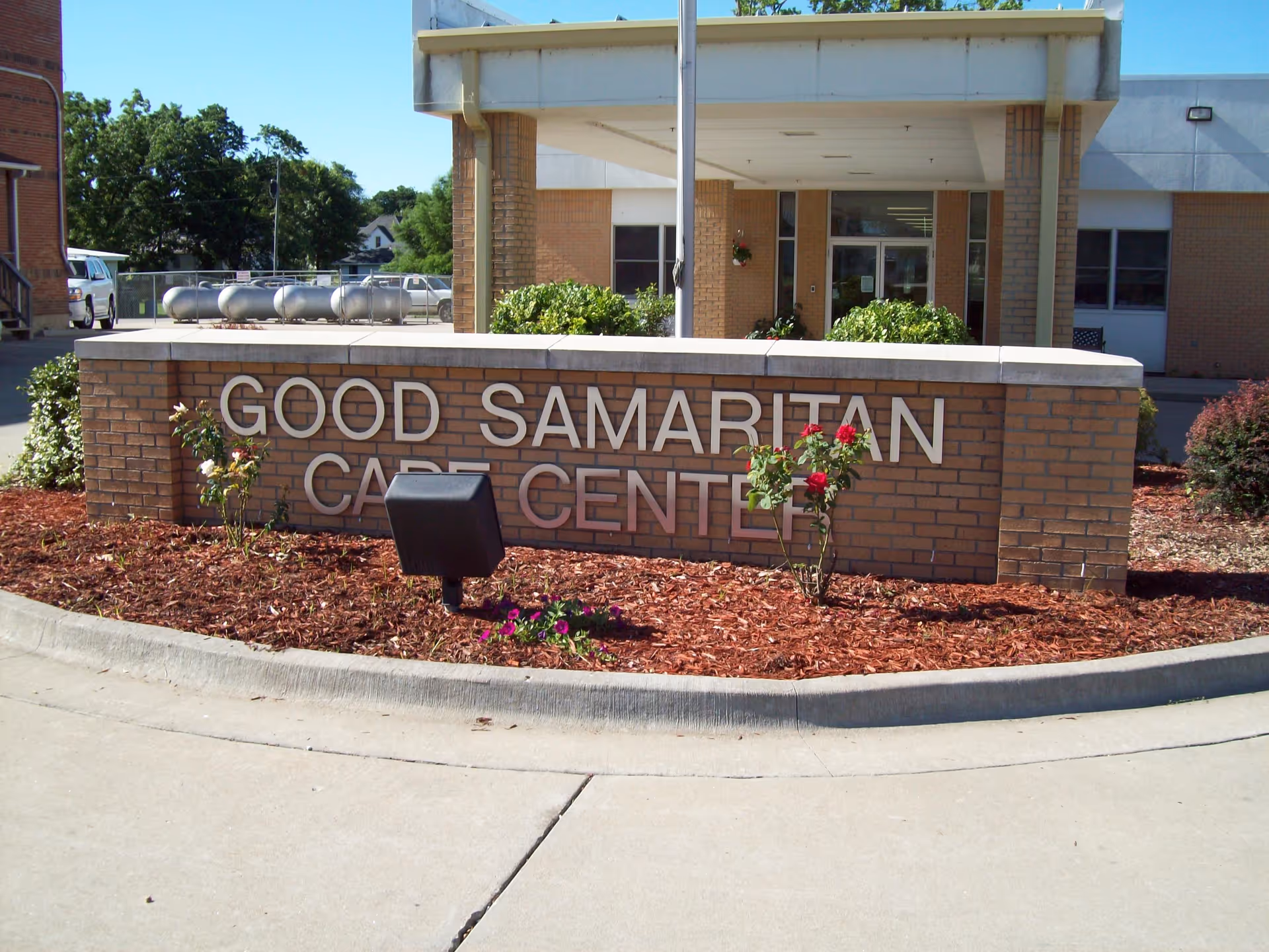 Exterior view of the entrance to Good Samaritan Care Center with a brick sign displaying the facility's name surrounded by mulch and flowers, and the building entrance visible in the background.