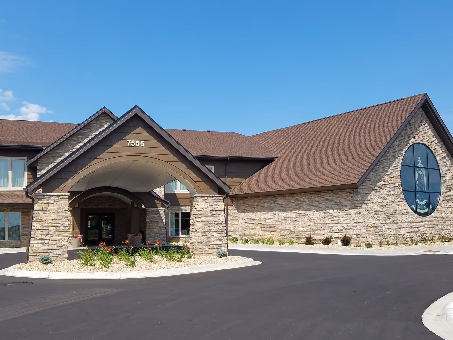 Exterior view of Saint Therese Senior Living of Woodbury building with stone facade, brown roof, and a covered entrance with the number 7555 above it. There is a circular driveway and some landscaping with flowers and plants in front of the entrance. A large round window with a religious figure is visible on the right side of the building.