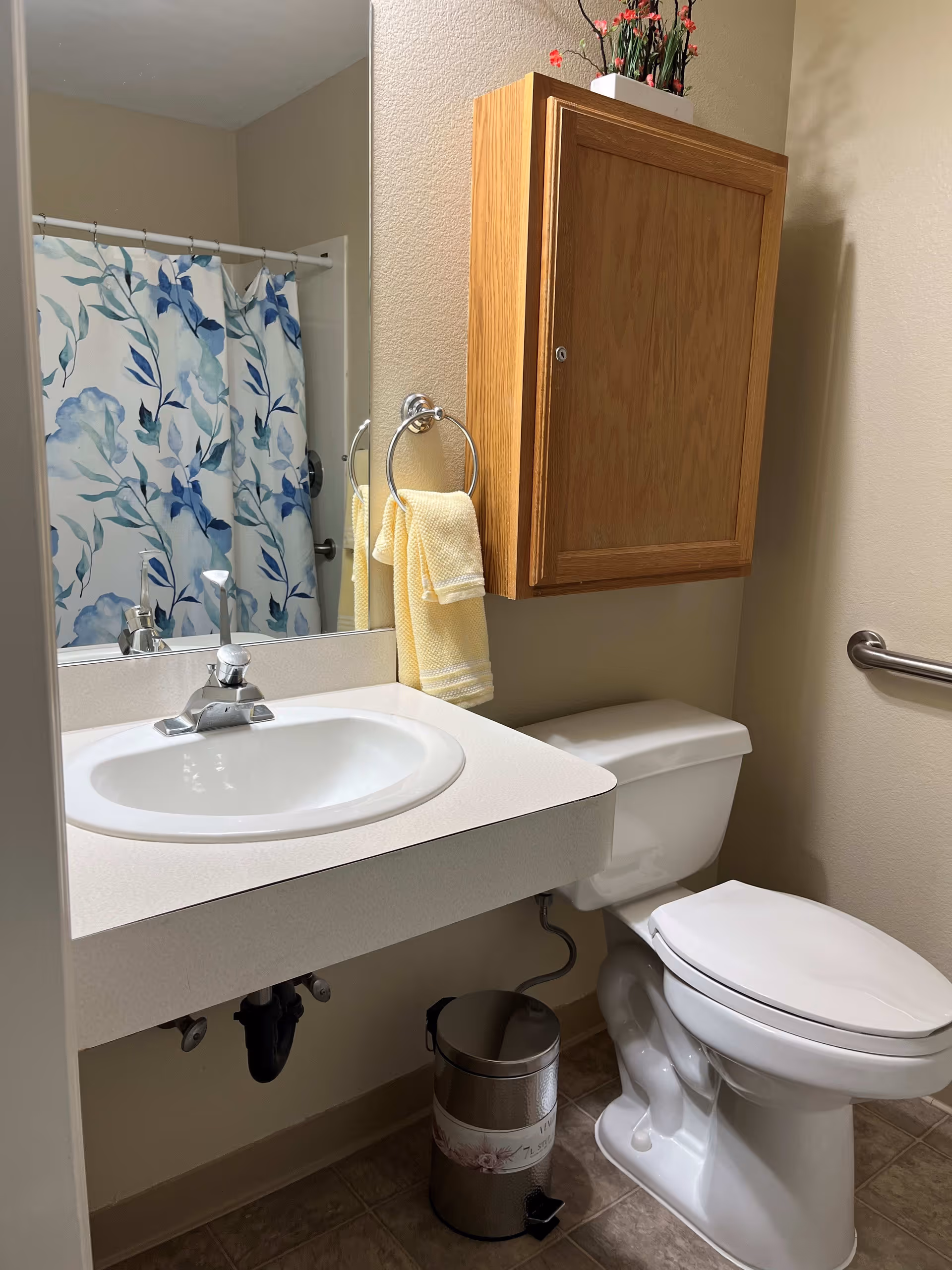 A clean bathroom featuring a white sink with a silver faucet, a wooden wall-mounted cabinet above a white toilet, a silver trash can on the floor, a towel ring holding two yellow towels, and a shower with a floral blue and white curtain visible in the mirror reflection.