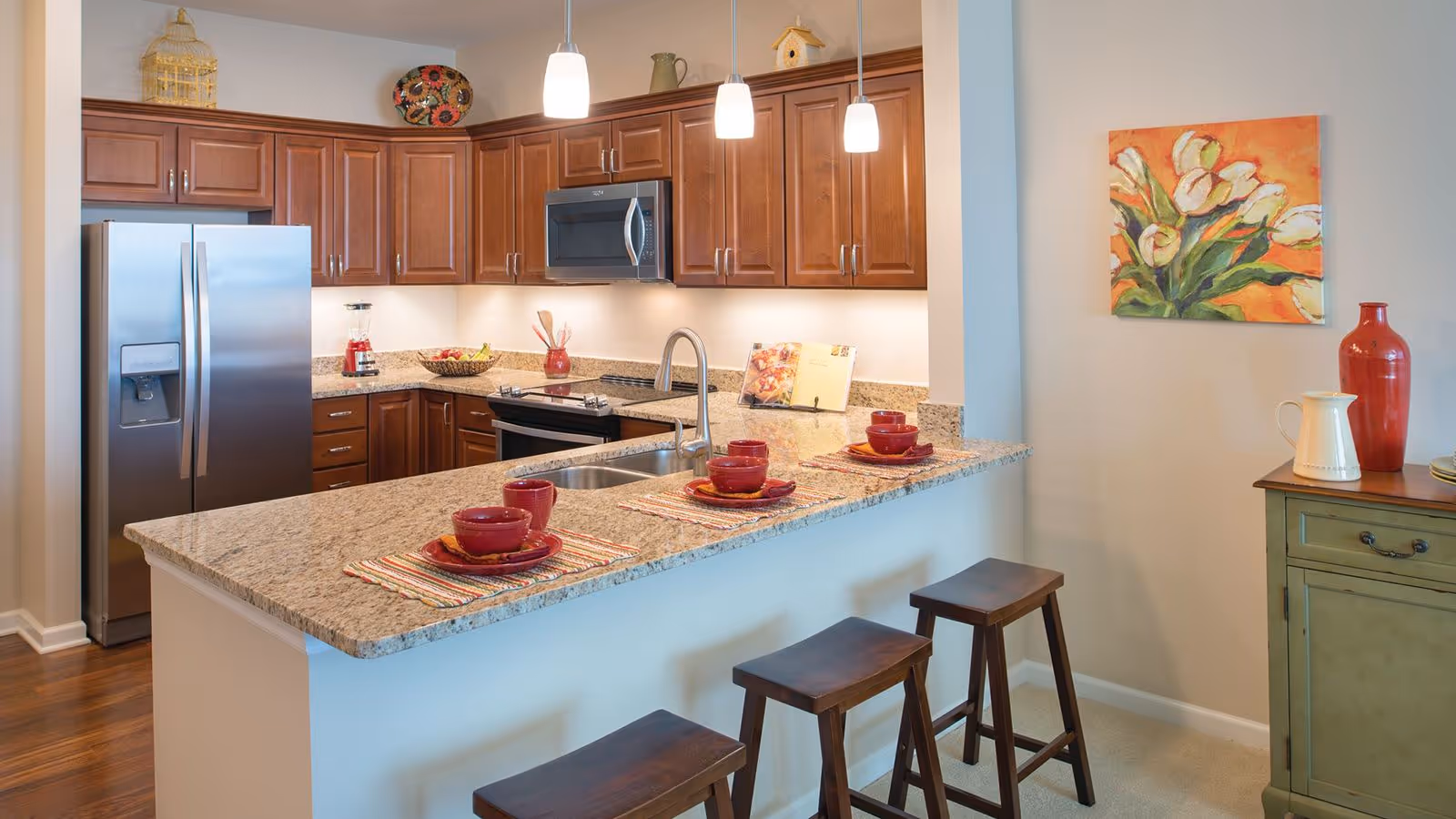 A modern kitchen with wooden cabinets, a stainless steel refrigerator, microwave, and oven. The kitchen features a granite countertop island with three wooden stools and place settings with red bowls and plates. There is a colorful painting of flowers on the wall and decorative items on a green side cabinet.