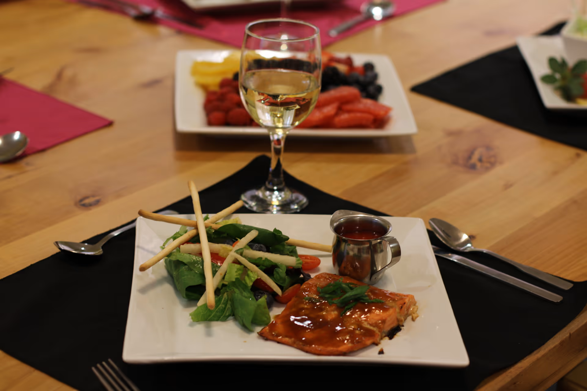 A plated meal on a wooden table featuring a piece of glazed salmon garnished with green herbs, a side salad with breadsticks, and a small metal container of sauce. A glass of white wine is placed behind the plate, and a plate of assorted fresh fruit is visible in the background. The table is set with black and red placemats and silverware.