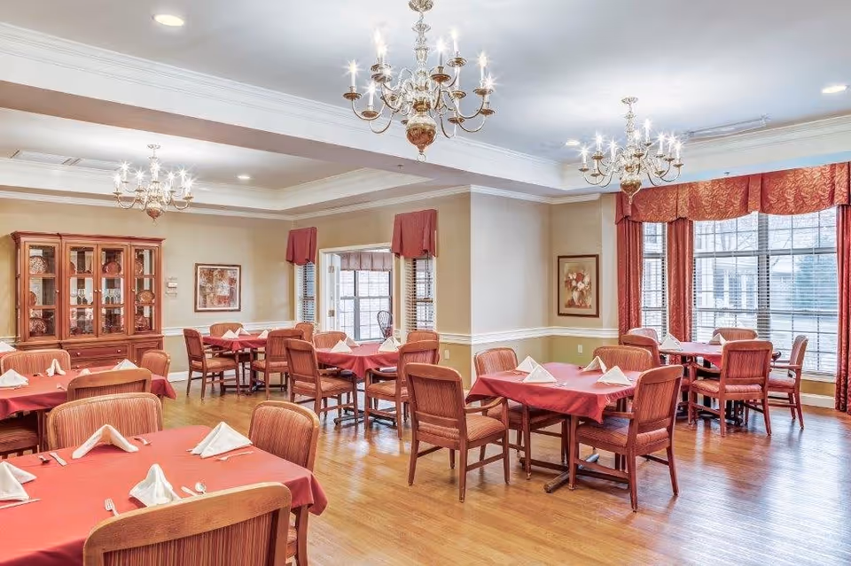 A bright dining room with multiple wooden tables covered with red tablecloths and neatly folded white napkins. The room features wooden chairs, large windows with red curtains, chandeliers hanging from the ceiling, and a wooden cabinet displaying decorative items.