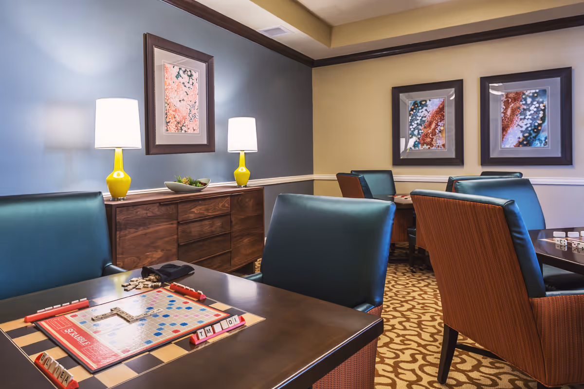 Well-lit common room with tables and green upholstered chairs set up for games, a Scrabble board on a table, framed artwork and lamps on a sideboard.