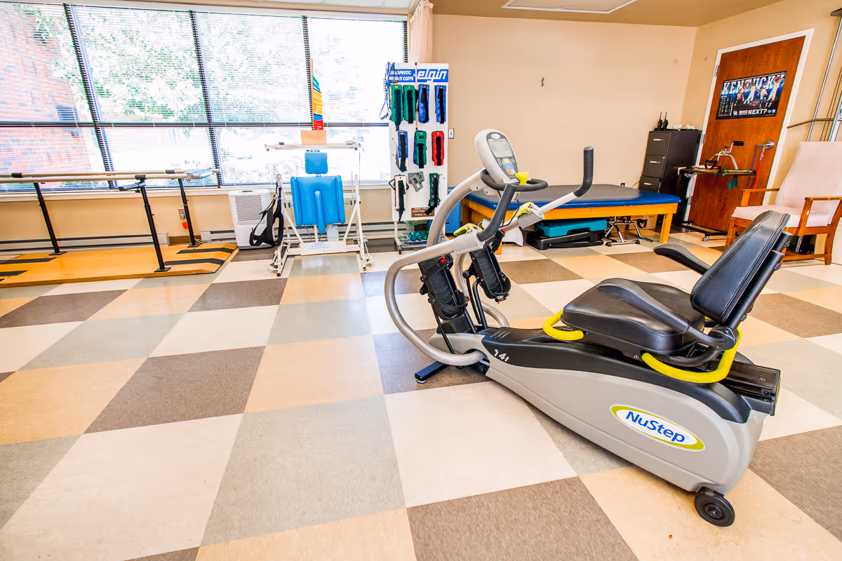 A rehabilitation and wellness room with exercise equipment including a NuStep recumbent cross trainer, parallel bars for walking support, a padded therapy table, and various physical therapy tools. The room has large windows with blinds, checkered flooring, and a chair near a wooden door with a Kentucky poster.
