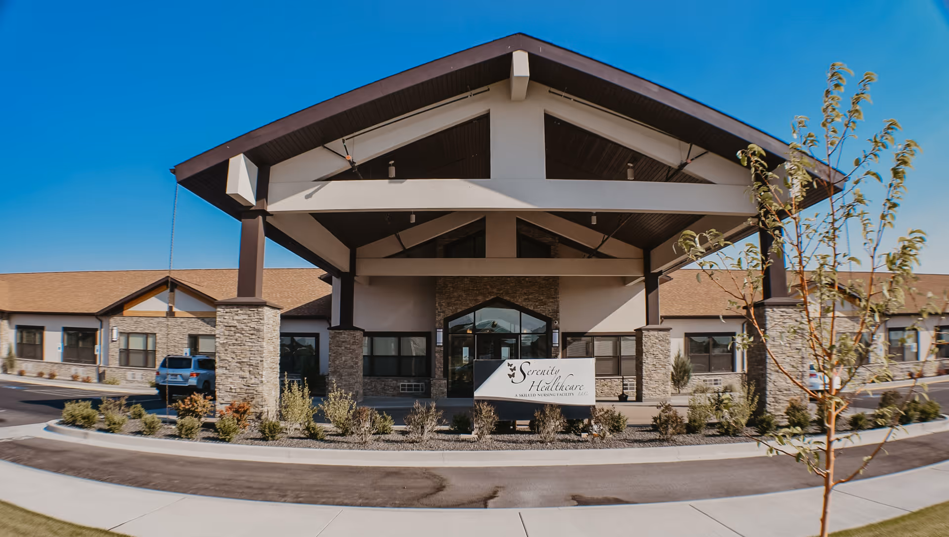 Front exterior view of Serenity Healthcare, a single-story building with stone pillars and a covered entrance, surrounded by landscaping and a clear blue sky.