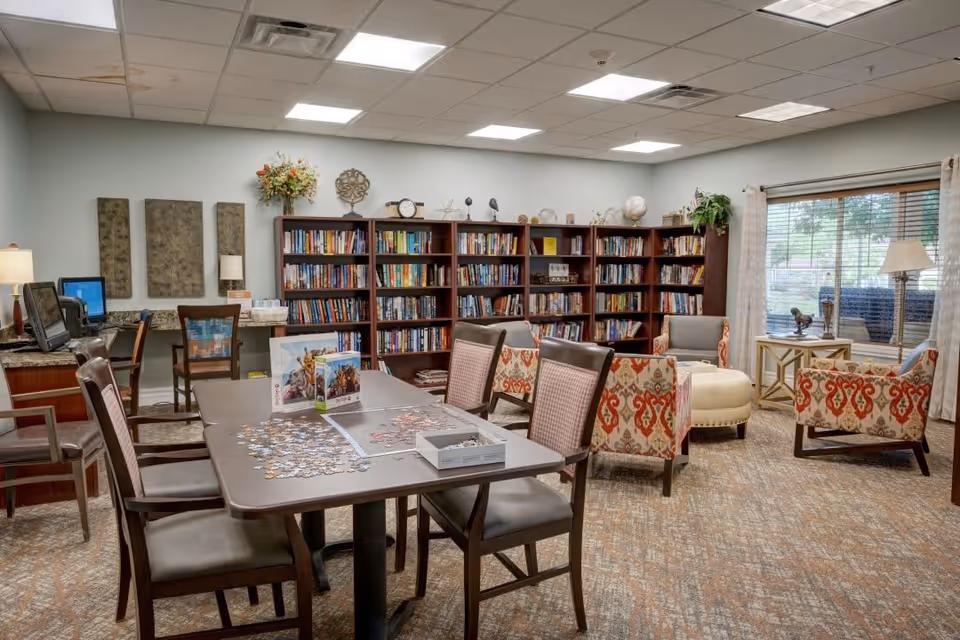A cozy common area with a large bookshelf filled with books, several chairs with patterned upholstery, a round ottoman, and a table with a partially completed jigsaw puzzle. The room has a window with curtains, a lamp on a side table, and a computer desk with chairs in the background.