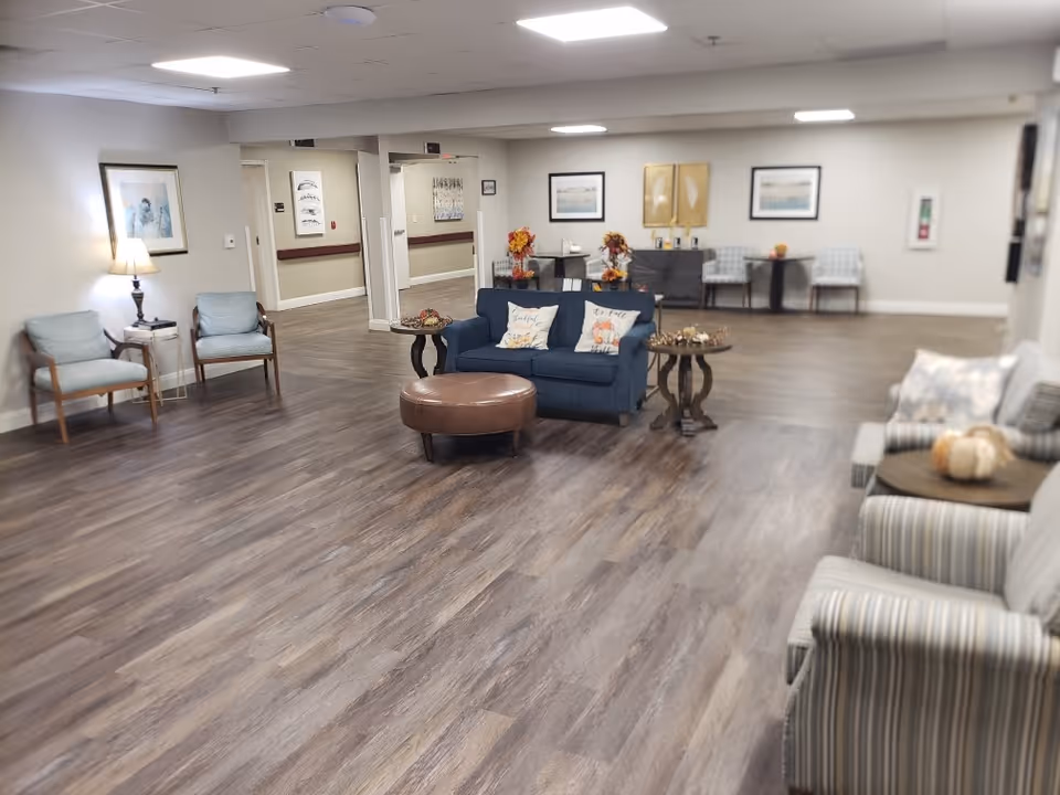 A spacious and well-lit common area in a senior living facility with wood flooring, multiple seating arrangements including armchairs, a blue loveseat with decorative pillows, small round tables, and framed artwork on the walls. The room has a cozy and welcoming atmosphere with soft lighting from ceiling fixtures and a table lamp.