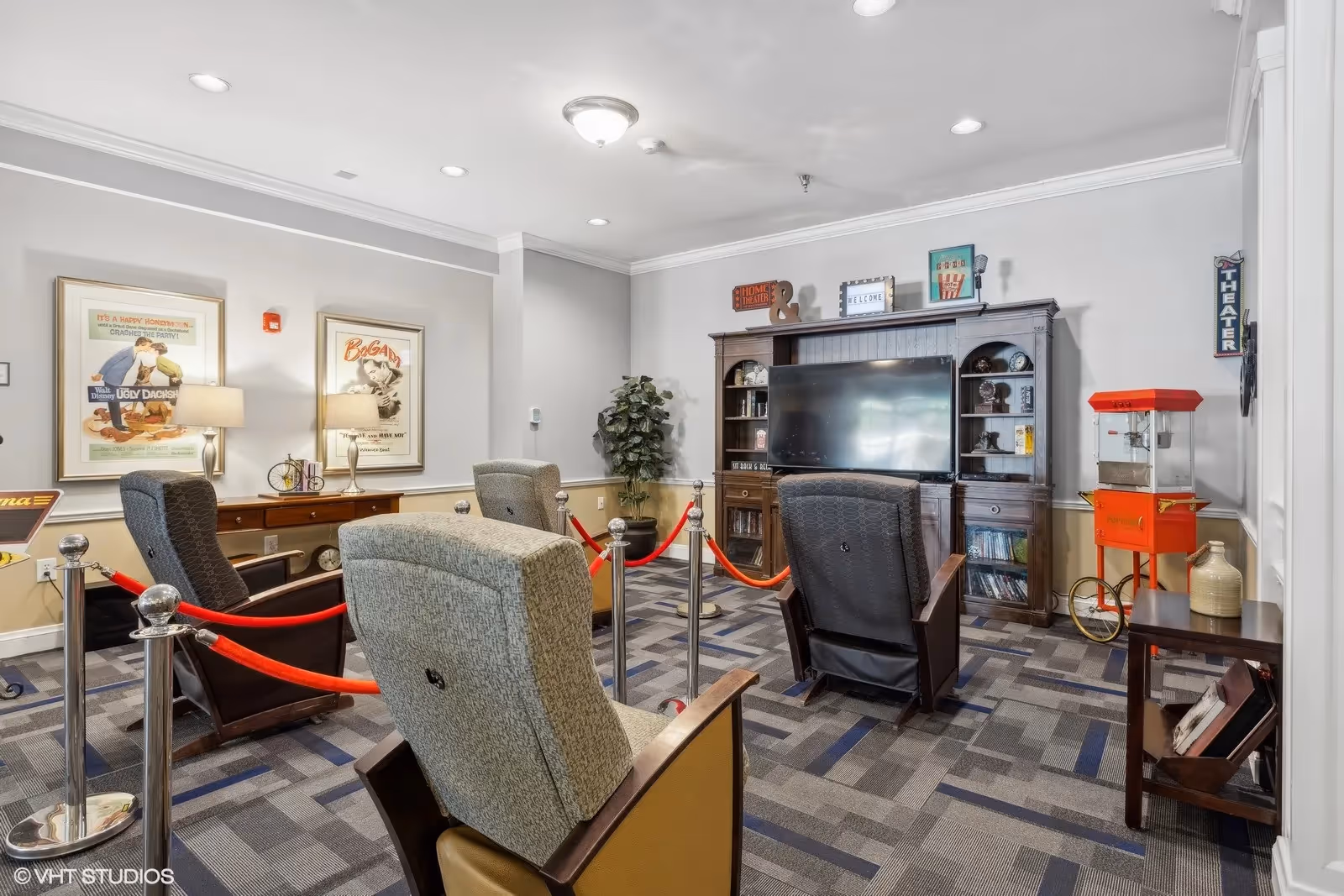 A cozy entertainment room with several comfortable armchairs arranged facing a large flat-screen TV mounted on a wooden entertainment center. The room features movie-themed decor including vintage movie posters on the wall, a popcorn machine, and a sign that says 'THEATER'. The floor is carpeted with a patterned design, and red velvet ropes separate the seating area.