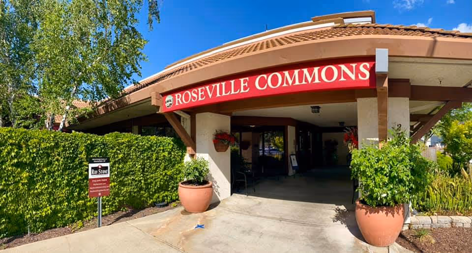Front entrance of Roseville Commons senior living with a covered porte-cochere, potted plants, and a red sign reading 'ROSEVILLE COMMONS'.