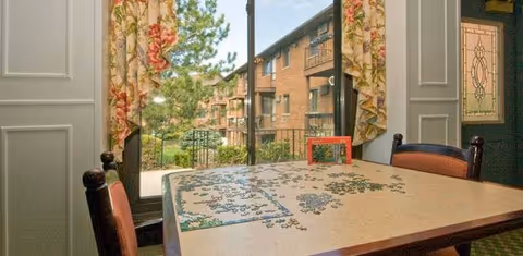 A table with a partially completed jigsaw puzzle sits in front of a large window with floral curtains. Outside the window, there is a view of a brick building with balconies and greenery. Two chairs are positioned at the table, and a door with decorative glass is visible on the right side.