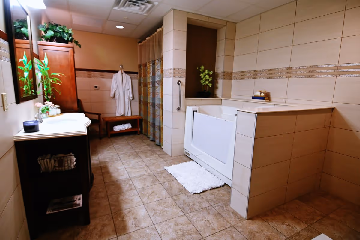 A spacious bathroom with beige tiled walls and floor. There is a white walk-in bathtub with a grab bar, a white bath mat in front of it, and a small plant on the tub ledge. On the left side, there is a dark wooden vanity with a white countertop, a sink, and a mirror above it. Next to the vanity is a wooden cabinet with green plants on top. In the background, a white bathrobe hangs on a hook above a wooden bench, and a shower area with a patterned curtain is visible.