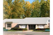 Single-story brick senior living building with a covered central entrance, shrubs in front and trees behind.