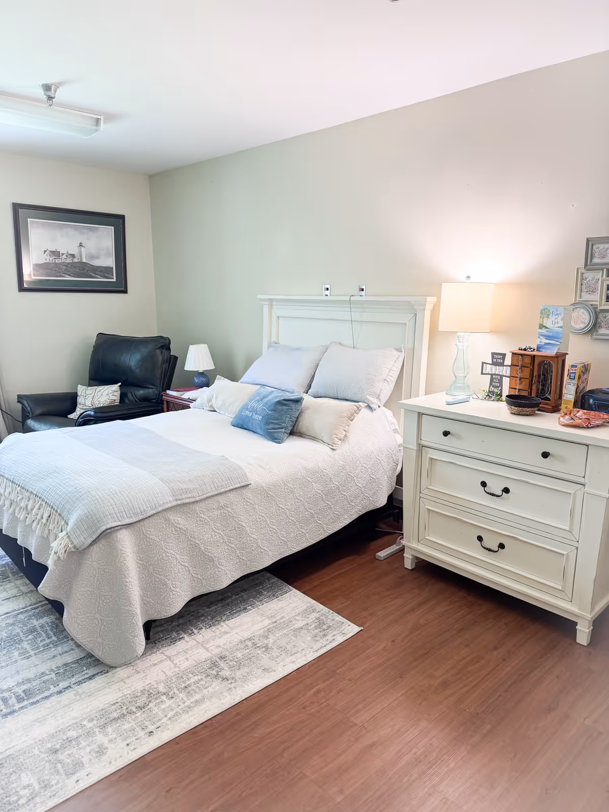 A cozy bedroom in an assisted living facility featuring a neatly made bed with white and light blue bedding, a white wooden headboard, and multiple pillows. To the right of the bed is a white dresser with three drawers, a table lamp, and various decorative items. To the left of the bed is a black leather armchair with a small side table and a lamp. A framed picture hangs on the wall above the armchair. The room has wooden flooring and a light-colored area rug partially under the bed.