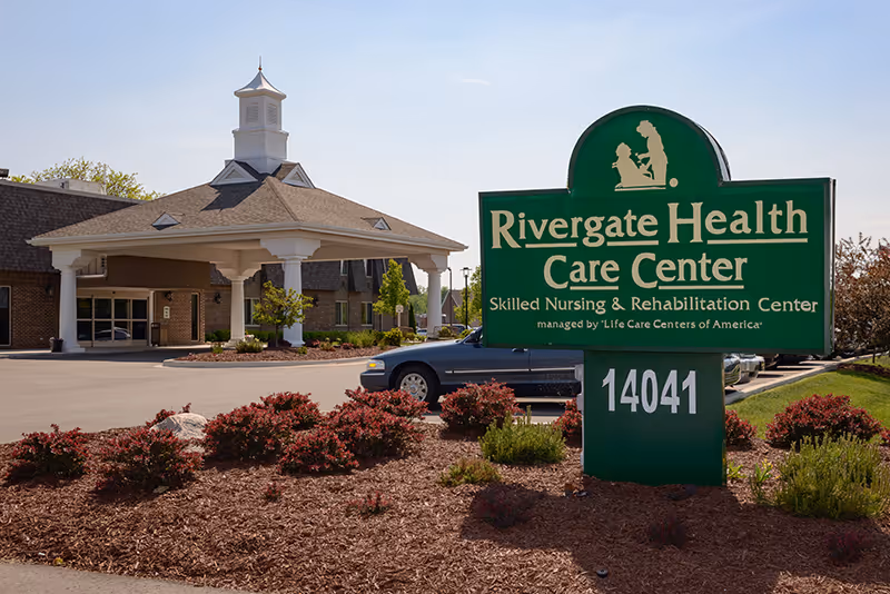 Exterior view of Rivergate Health Care Center, a skilled nursing and rehabilitation center, showing the main entrance with a covered drop-off area, landscaped bushes, and a green sign with the facility name and address 14041.
