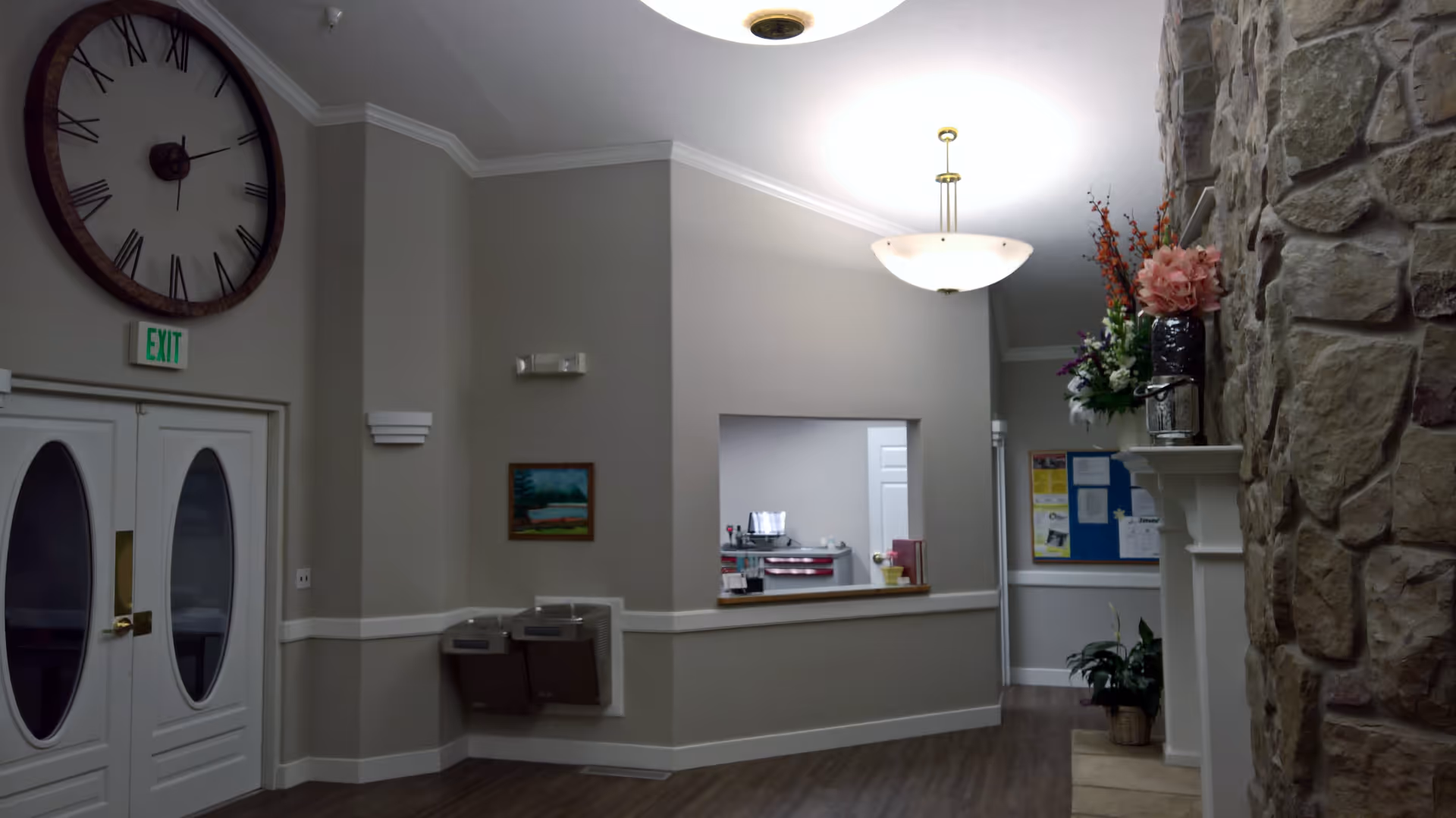 Interior view of a senior living facility hallway with a large wall clock above double doors, an exit sign, two water fountains, a reception window, a bulletin board, and a stone wall with floral arrangements on a mantel.