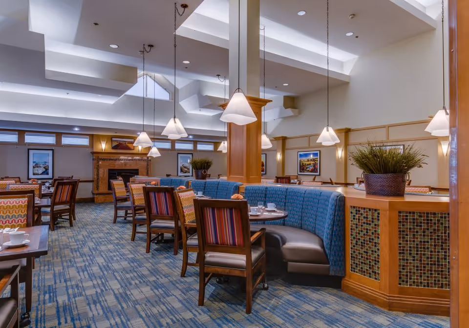 A spacious dining area in a senior living facility featuring multiple wooden tables and chairs with colorful upholstery. There is a curved blue booth seating area around a wooden partition with mosaic tile accents. Pendant lights hang from the ceiling, and framed artwork decorates the walls. The room has a patterned blue carpet and a fireplace in the background.