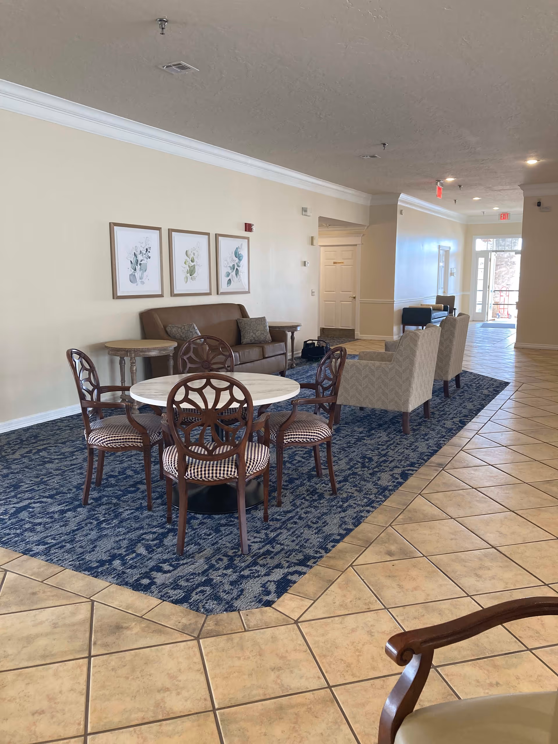 A spacious and well-lit common area with beige tiled flooring and a blue patterned rug. The area features a round table with four wooden chairs that have patterned cushions. Behind the table is a brown leather sofa with two side tables and three framed botanical prints on the wall above it. Further back, there are two upholstered armchairs and additional seating near a glass door letting in natural light.