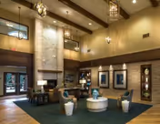 Spacious and well-lit senior living facility common area with high ceilings, a large stone fireplace, comfortable seating including armchairs and a round coffee table, decorative wall niches with art pieces, and large windows near the entrance letting in natural light.