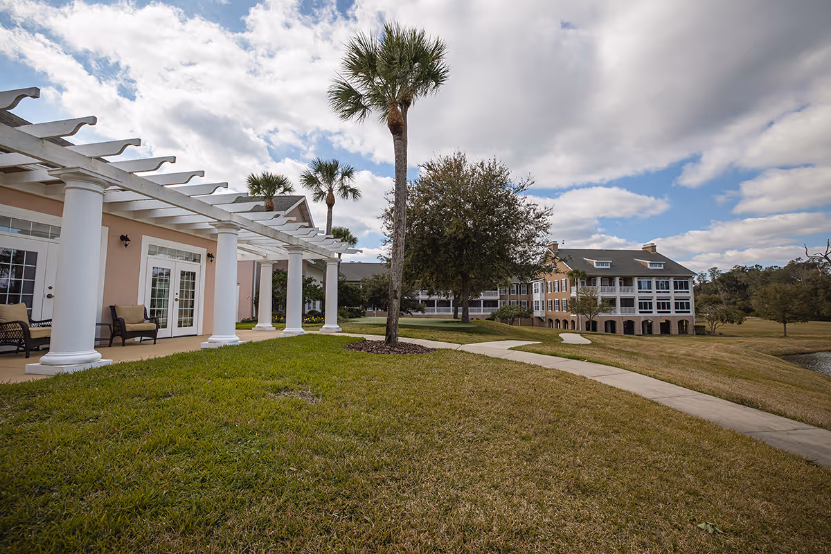 Exterior view of a senior living facility featuring a white-columned pergola, palm trees, a grassy lawn with a winding sidewalk, and a multi-story residential building under a partly cloudy sky.