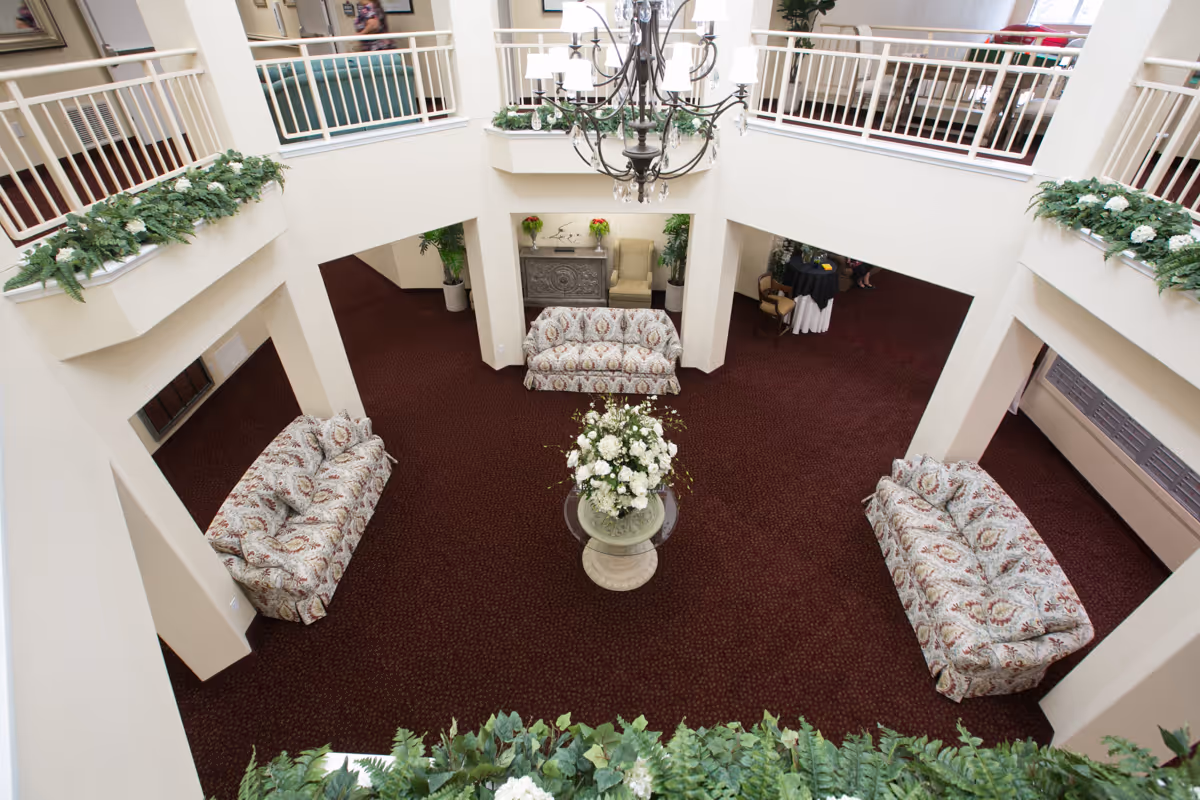 View from above of a spacious, carpeted common area in a retirement living facility with three patterned sofas arranged around a central table holding a large floral arrangement. The area is surrounded by white railings with green plants and white flowers decorating the upper level. A chandelier hangs from the ceiling above the seating area.