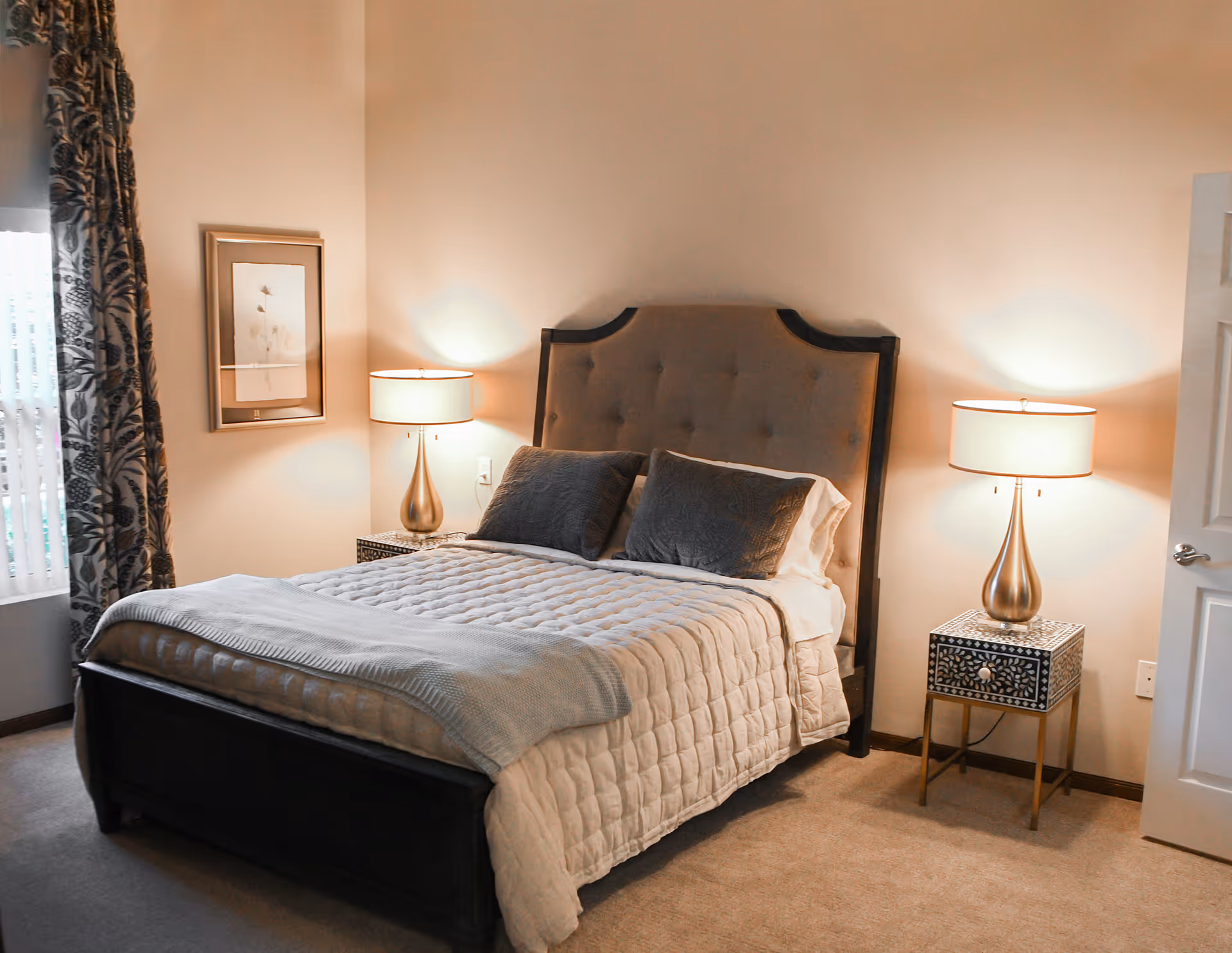 A neatly made bedroom with a tufted headboard bed, quilted bedding, two bedside lamps on decorative nightstands, patterned curtains, and framed wall art.
