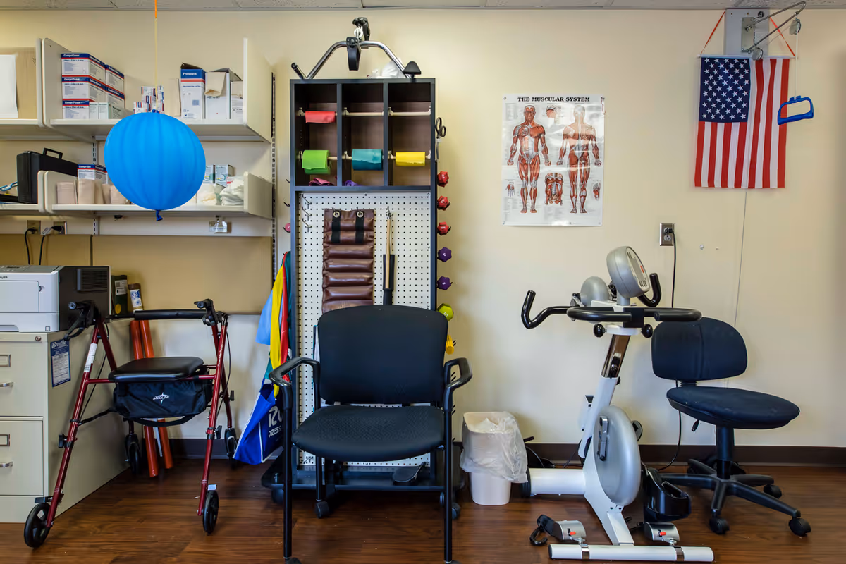 A small therapy or exercise room with a walker, two chairs, an exercise bike, and various therapy equipment including resistance bands and weights. There is a poster of the muscular system on the wall and an American flag hanging beside it. Shelves with medical supplies are visible on the left side.