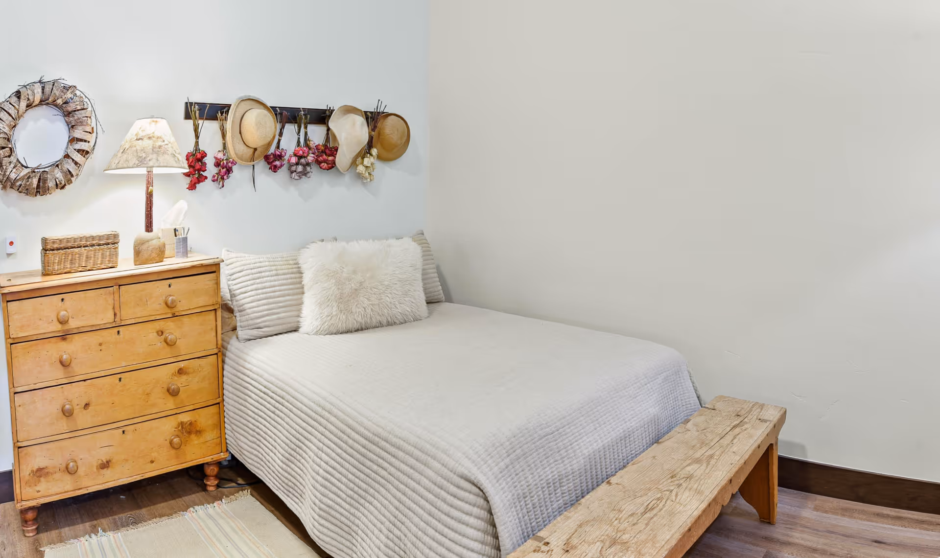 A simple bedroom featuring a bed with neutral bedding, a wooden dresser and bench, and hats and wall decor.