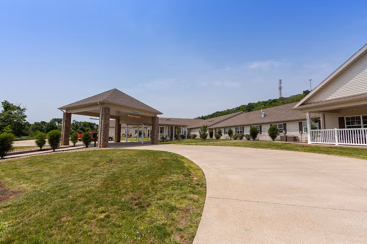 Exterior view of Belpre Landing Skilled Nursing & Rehabilitation facility showing a long, single-story building with a covered entrance driveway, surrounded by green grass and shrubs under a clear blue sky.