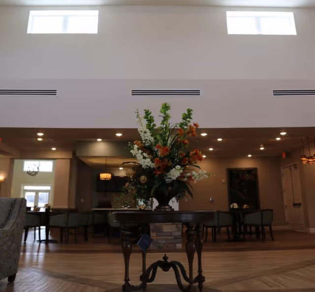 Interior view of a senior living facility common area with a round wooden table in the center holding a large floral arrangement. The room has high ceilings with two rectangular windows near the top, recessed lighting, and several chairs and tables in the background.