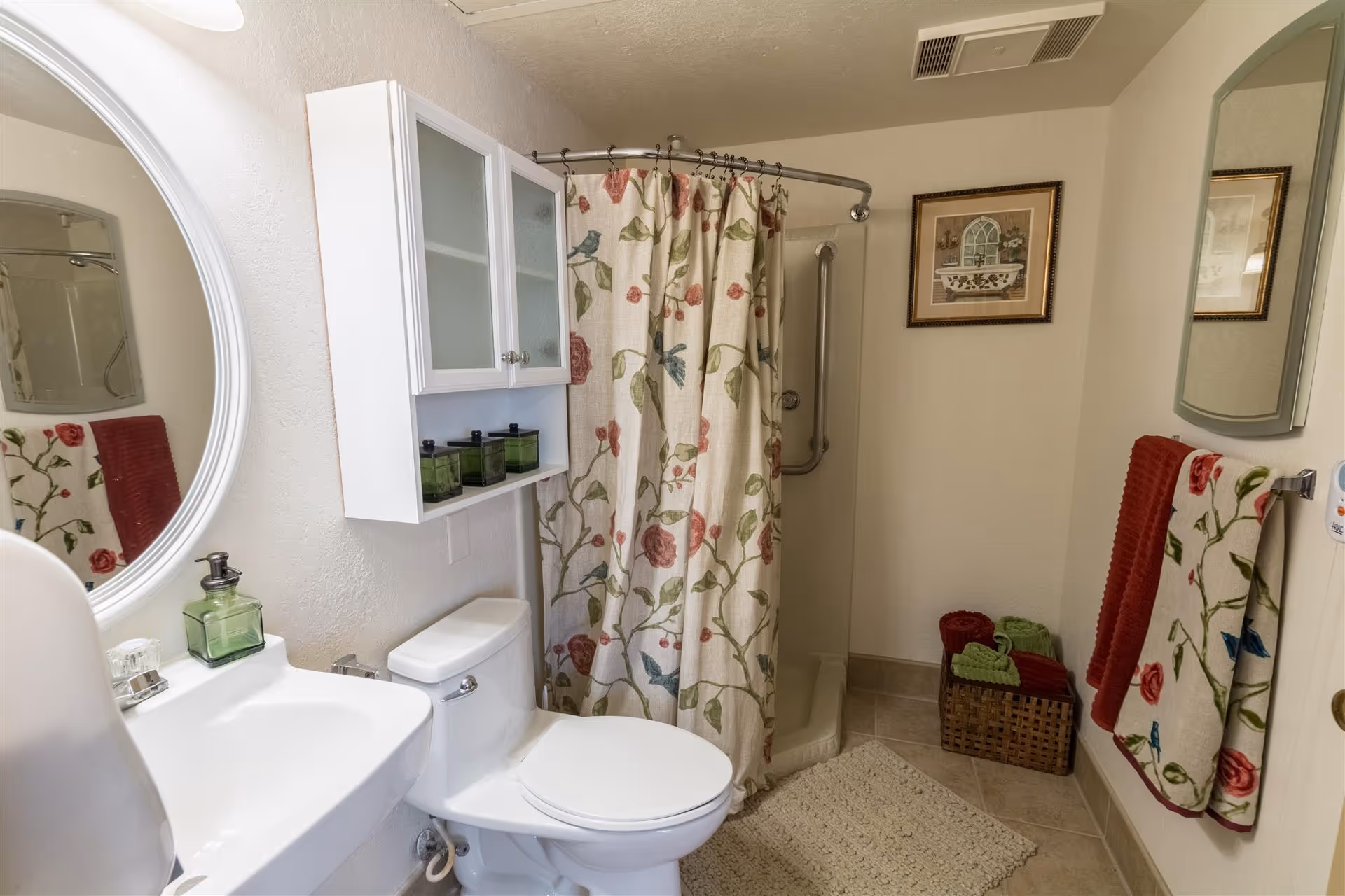 Bright, compact bathroom with a sink and round mirror, toilet, floral shower curtain over a walk-in shower, white wall cabinet, and matching floral towels.