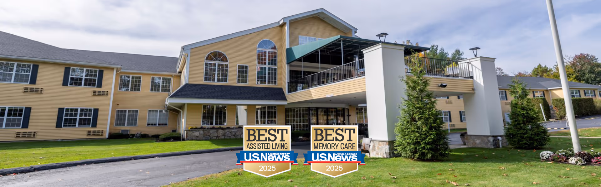 Front exterior of a yellow two-story senior living building with a covered entrance, balcony, landscaped lawn, and two 'Best Assisted Living / Best Memory Care US News 2025' signs on the driveway median.