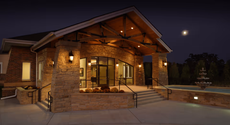 Night view of the entrance to a senior living facility with stone pillars, warm lighting, a covered porch, and a water fountain to the right under a dark sky with a visible moon.