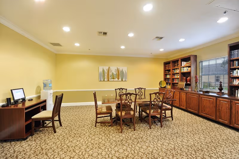 A well-lit room with yellow walls and patterned carpet featuring a wooden table surrounded by six chairs in the center. To the right, there is a large wooden bookshelf filled with books and decorative items beneath a window with blinds. On the left side, there is a small wooden desk with a chair and a water cooler next to it. A framed artwork with leaf designs hangs on the wall above the table.