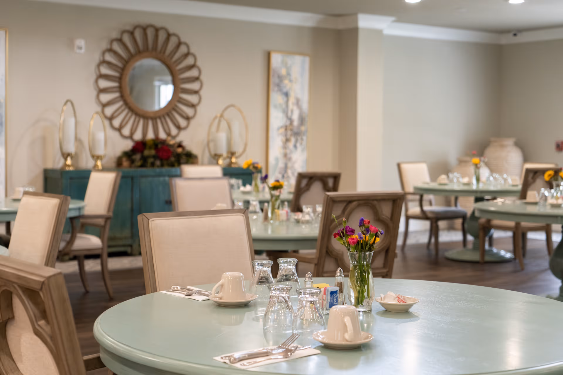 A dining room with round tables set with cups, glasses, silverware, and small vases of colorful flowers. The room has beige walls, cushioned chairs, a decorative mirror, and artwork on the walls.
